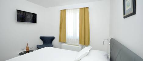 A hotel room with a bed, a chair, a small table with a bottle and glasses, a window with curtains, and a framed picture on the wall.
