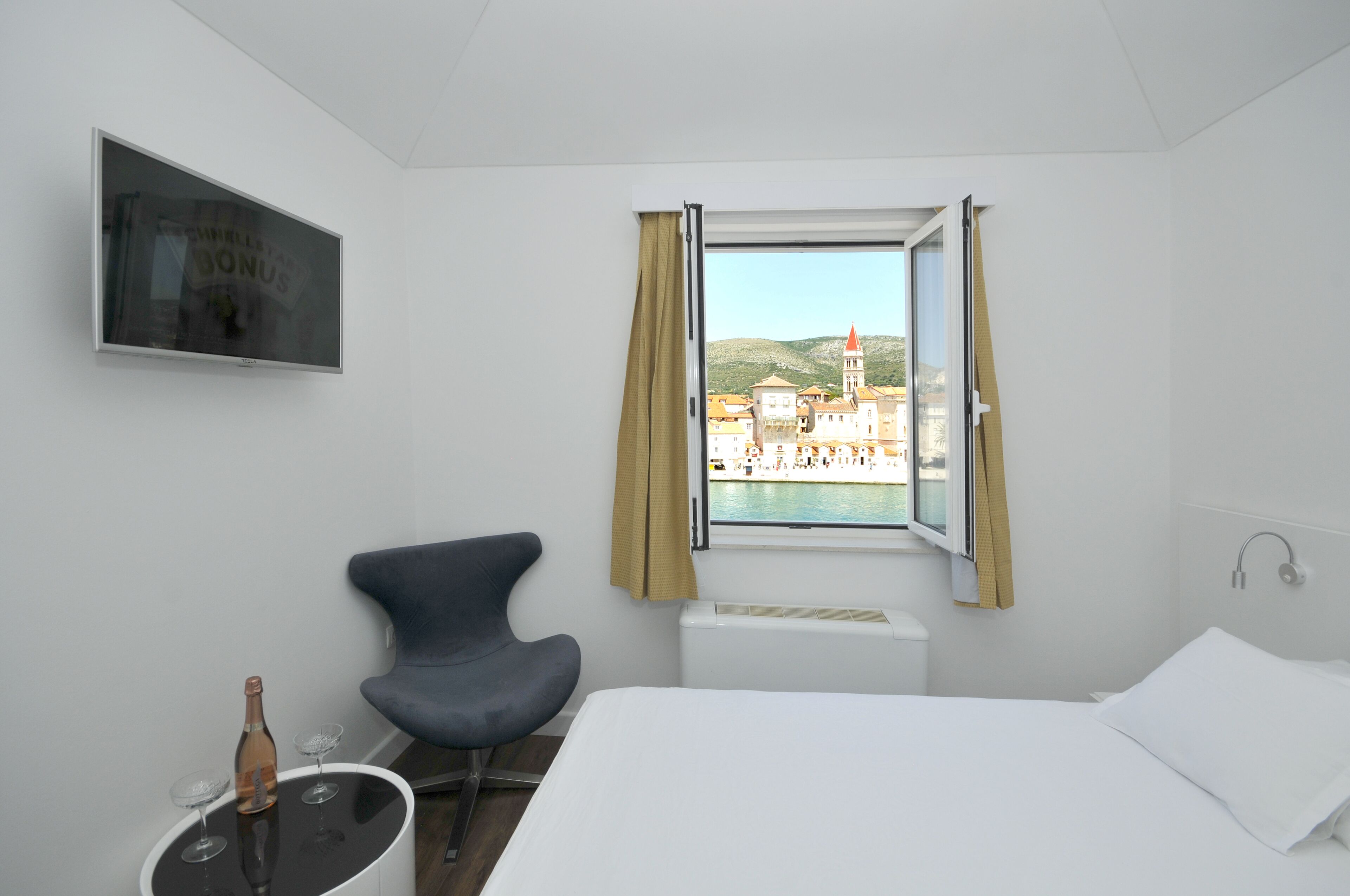 Una habitación de hotel con una cama, una silla, una mesita con una botella y vasos, un televisor en la pared y una ventana con cortinas.