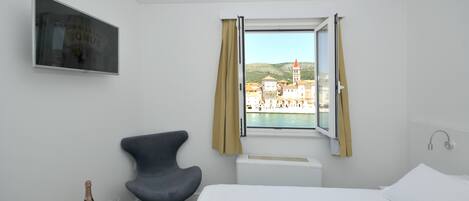 Una habitación de hotel con una cama, una silla, una mesita con una botella y vasos, un televisor en la pared y una ventana con cortinas.