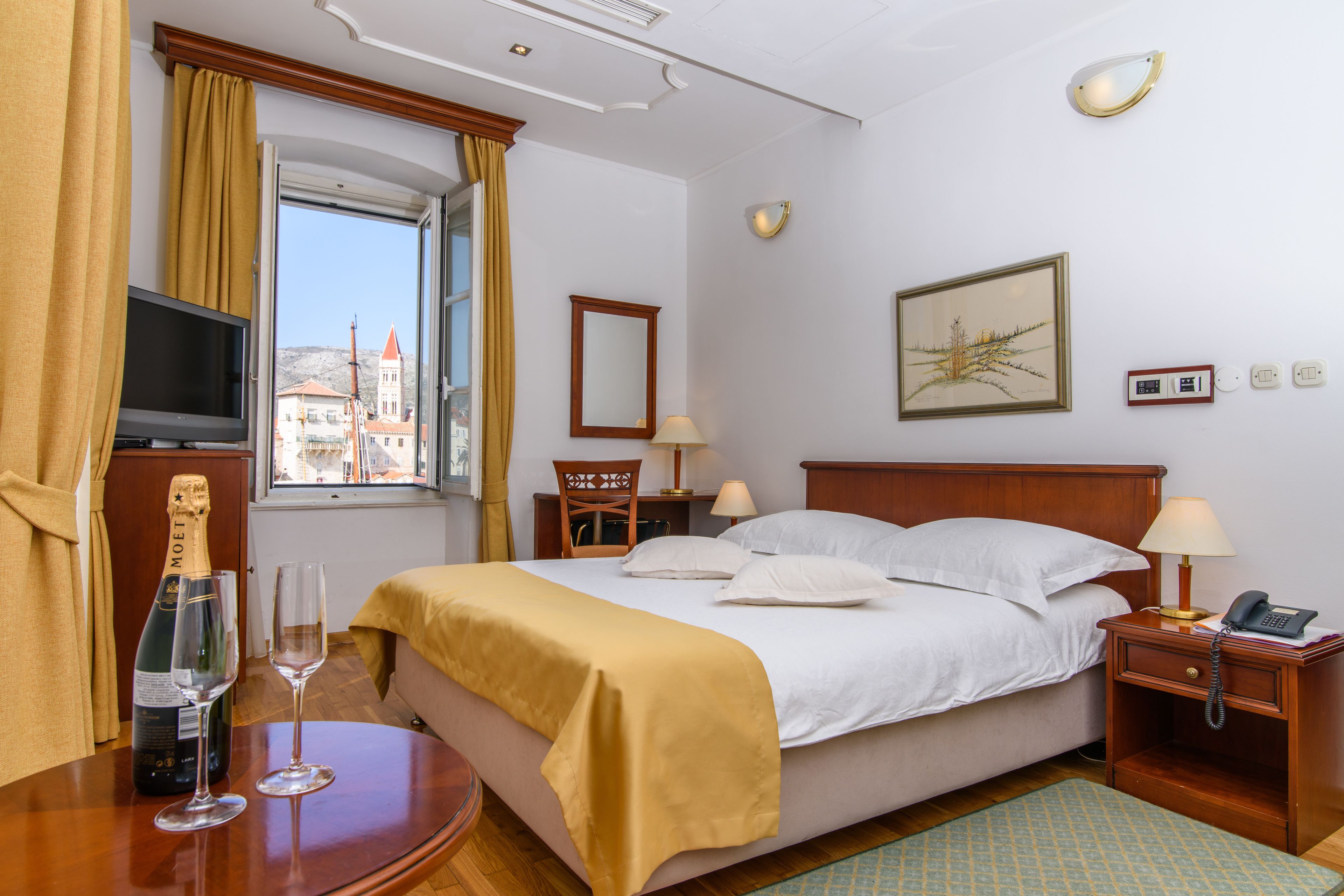 Habitación de hotel con cama, mesitas de noche, televisor, ventana con cortinas, un cuadro en la pared y una botella de champán con copas.