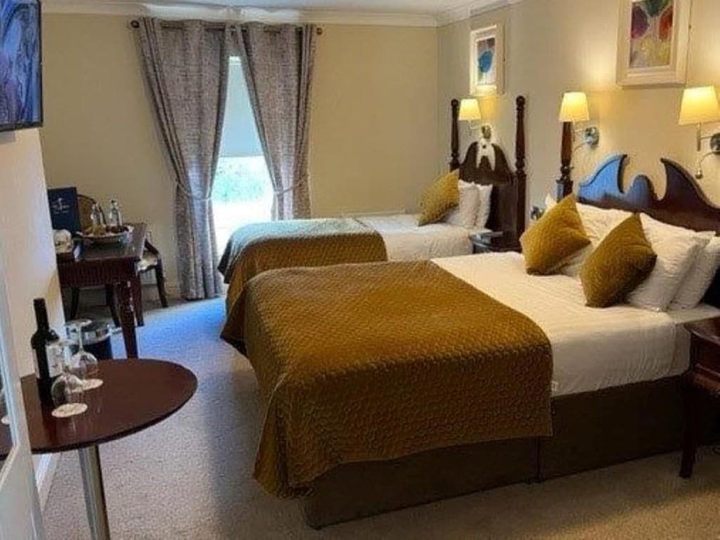 A hotel room with two beds, a small round table with a bottle and glasses, and a view of the outdoors through the window.