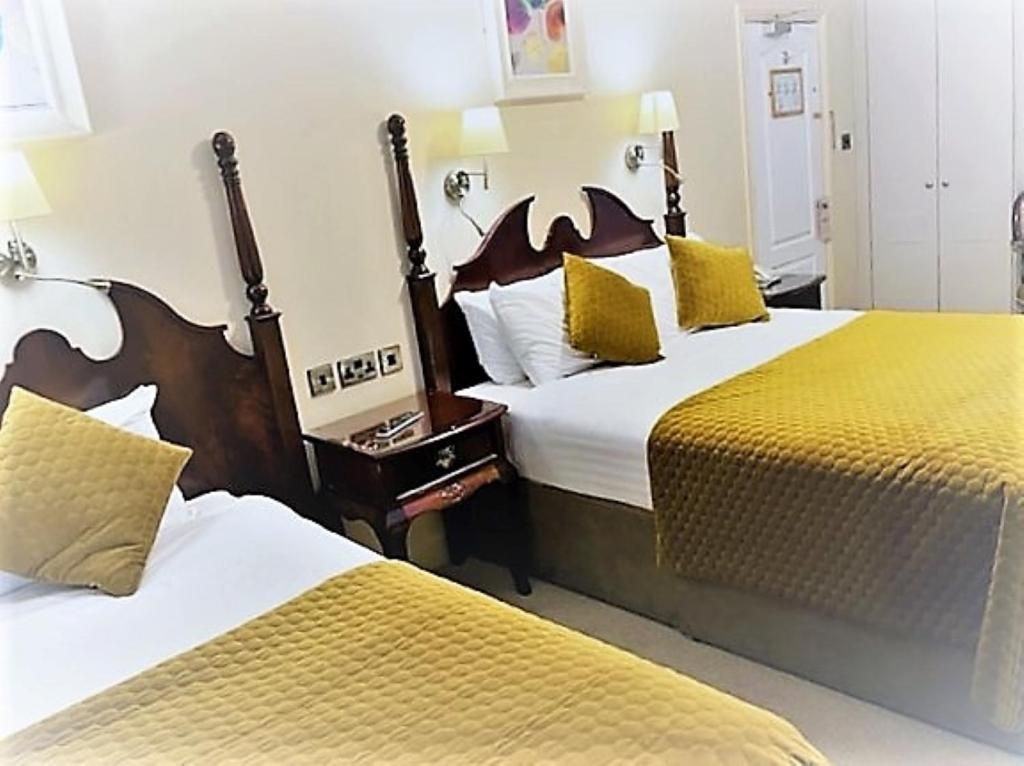 A hotel room with two beds, each with a wooden headboard, white linens, and yellow pillows. There is a nightstand with a lamp between the beds.