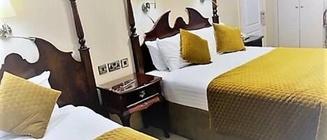 A hotel room with two beds, each with a wooden headboard, white linens, and yellow pillows. There is a nightstand with a lamp between the beds.