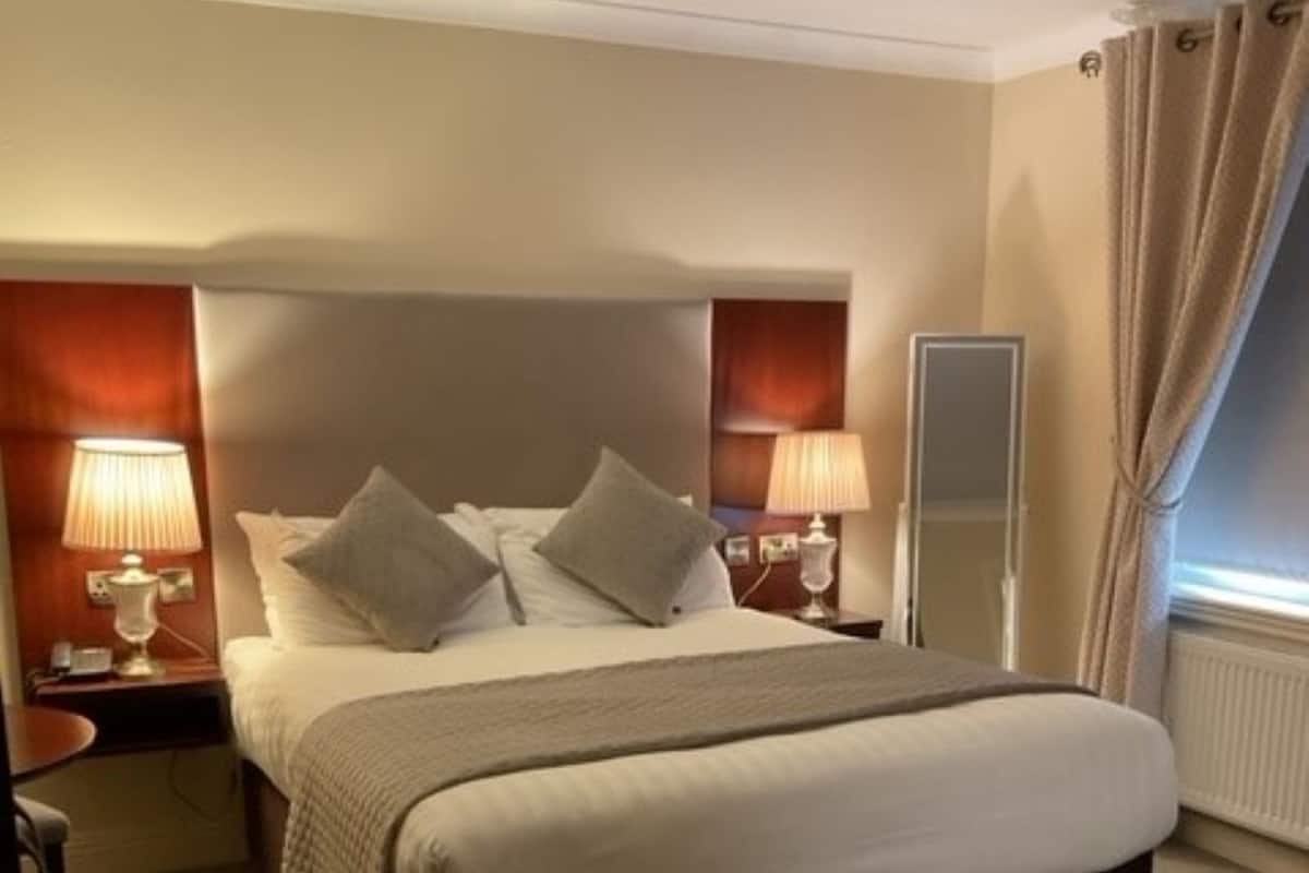 A hotel room with a large bed, two bedside tables with lamps, a mirror, and a view of the outdoors through a window with curtains.