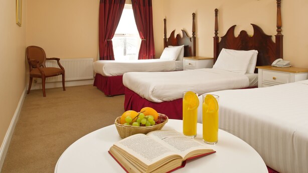 A hotel room with two beds, a chair, a bedside table with a lamp, a window with red curtains, and a table with a book and fruit. - Shamrock Inn Hotel (Lahinch)