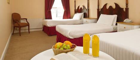 A hotel room with two beds, a chair, a bedside table with a lamp, a window with red curtains, and a table with a book and fruit.