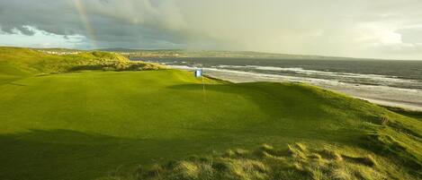 A golf course with a flagstick on the green, a view of the ocean, and a cloudy sky.