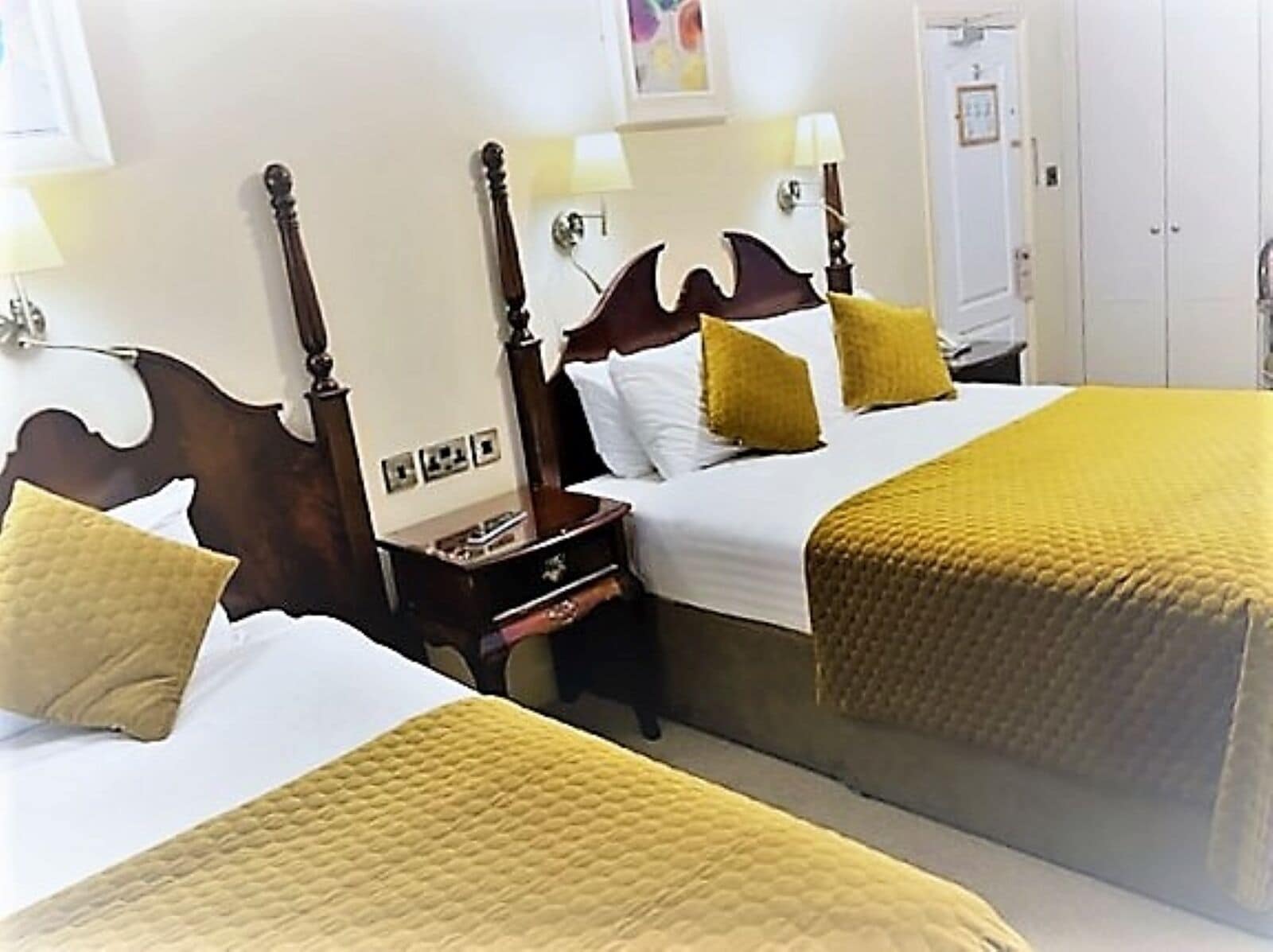 Une chambre d’hôtel avec deux lits, chacun doté d’une tête de lit en bois, de draps blancs et d’un plaid jaune. On y trouve des tables de chevet avec des lampes, ainsi qu’une petite table de nuit avec un livre et un téléphone.