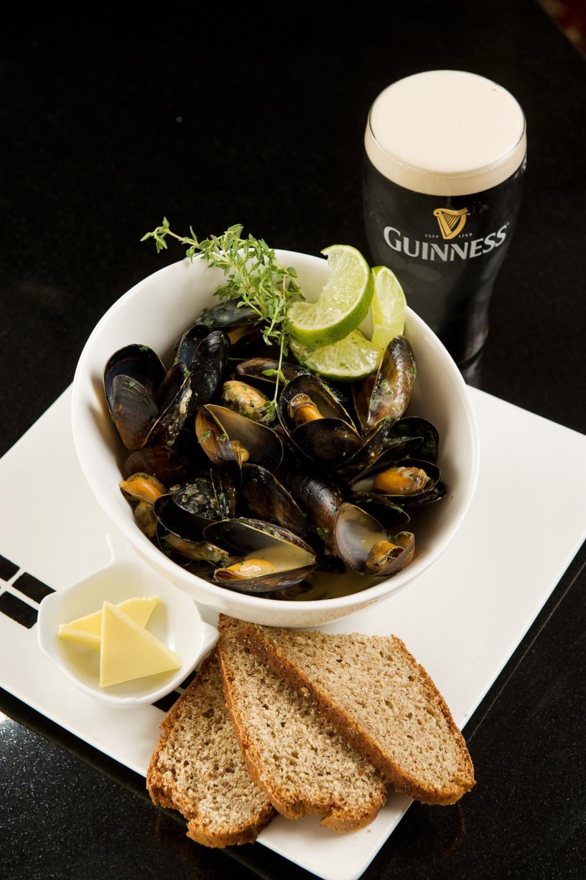 A bowl of mussels with a garnish, a glass of Guinness beer, a small bowl of butter, and slices of bread on a table.