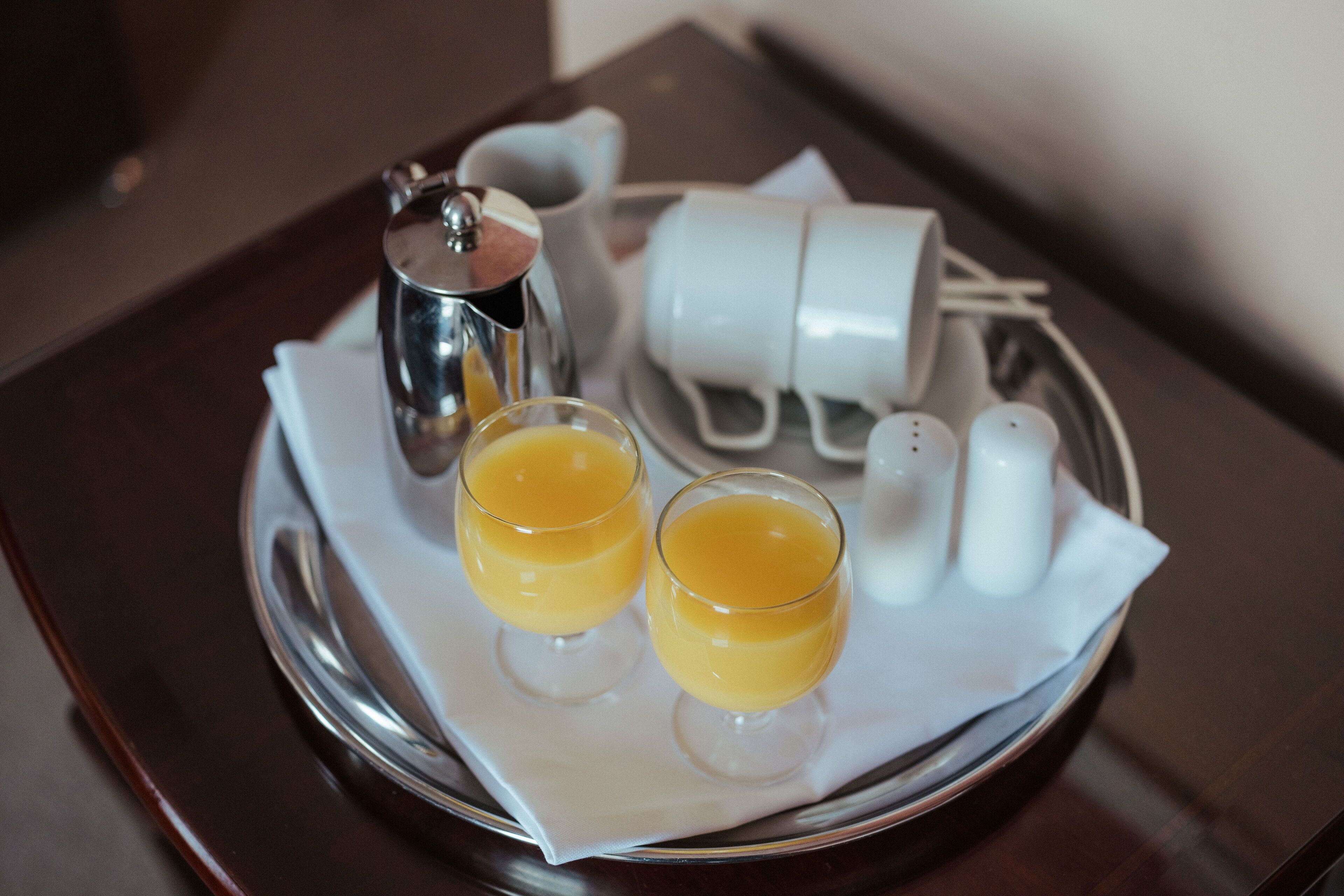 Una bandeja con dos vasos de jugo de naranja, una cafetera de plata y un juego de tazas y platones blancos.