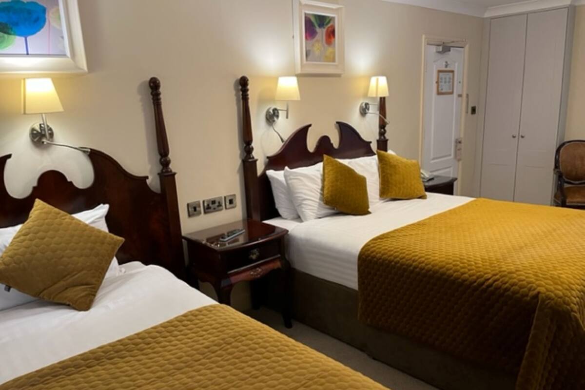 Habitación de hotel con dos camas, cada una con cabecero de madera, ropa de cama blanca y colcha color mostaza. Hay lámparas de noche y una mesita de noche con un libro.
