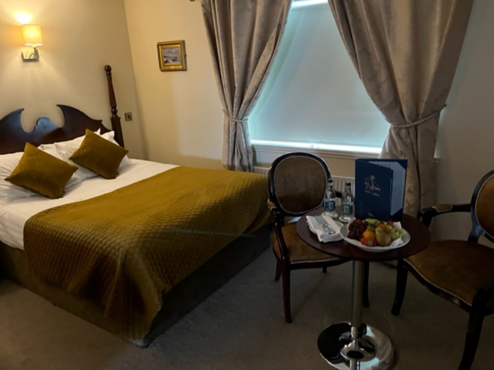 A hotel room with a bed, two chairs, a small table with a plate of fruit, a lamp, and a window with curtains.