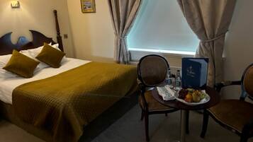 A hotel room with a bed, two chairs, a small table with a plate of fruit, a lamp, and a window with curtains.