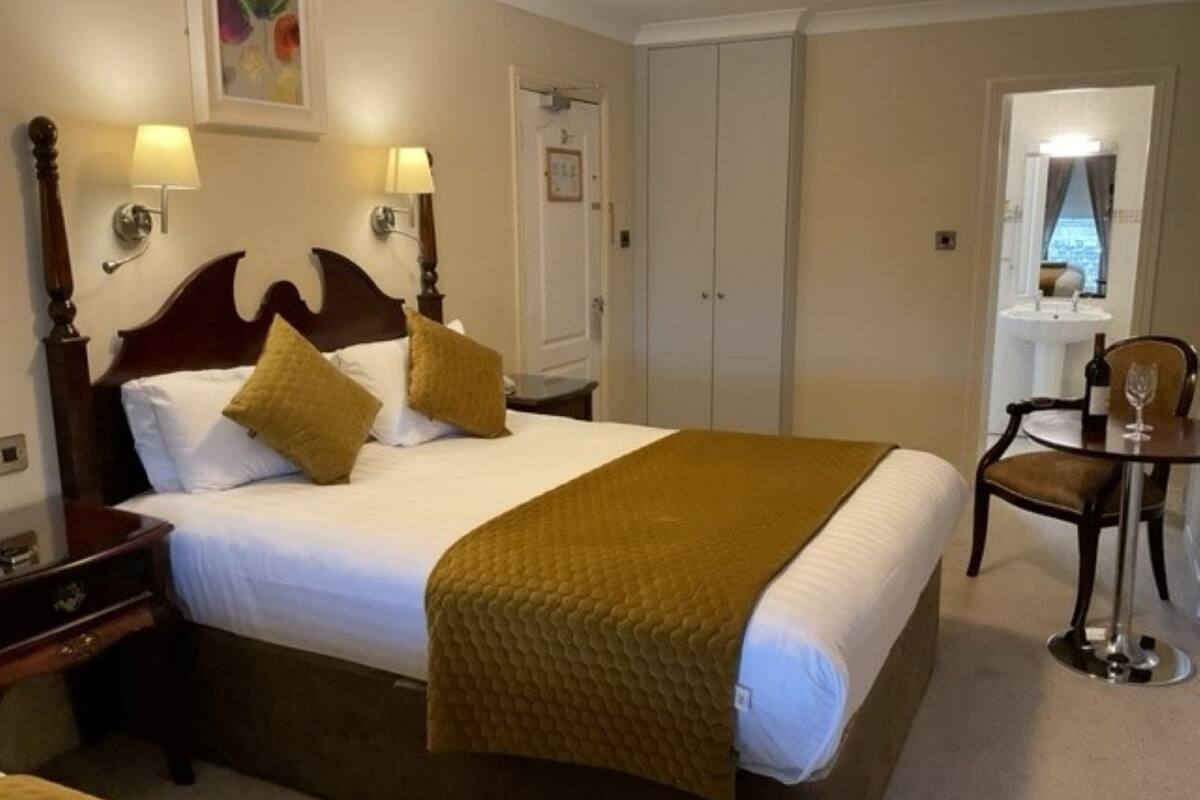 Habitación de hotel con una cama grande, dos lámparas de noche, un cabecero de madera, un plaid marrón, una mesita con una silla y vistas a un paisaje urbano a través de una ventana.
