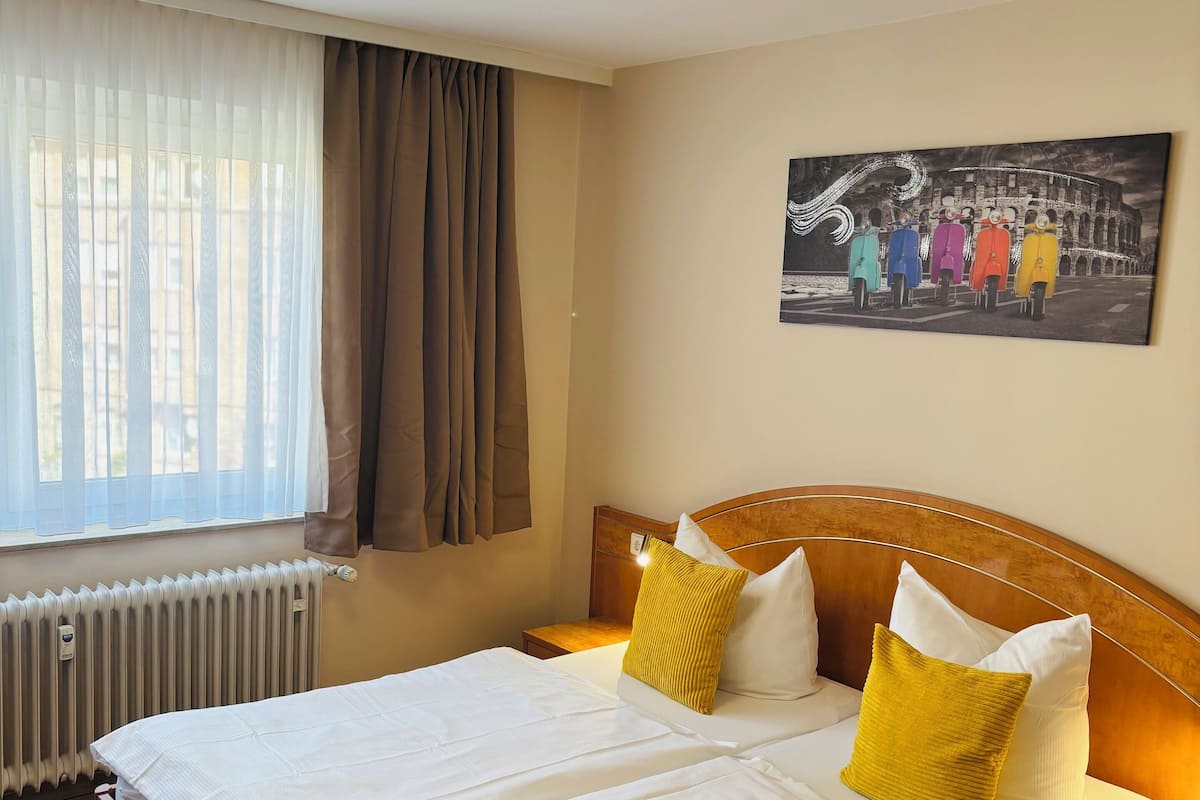 A hotel room with a large bed, a headboard, a bedside table, a nightstand, a window with curtains, a radiator, and a wall-mounted picture.