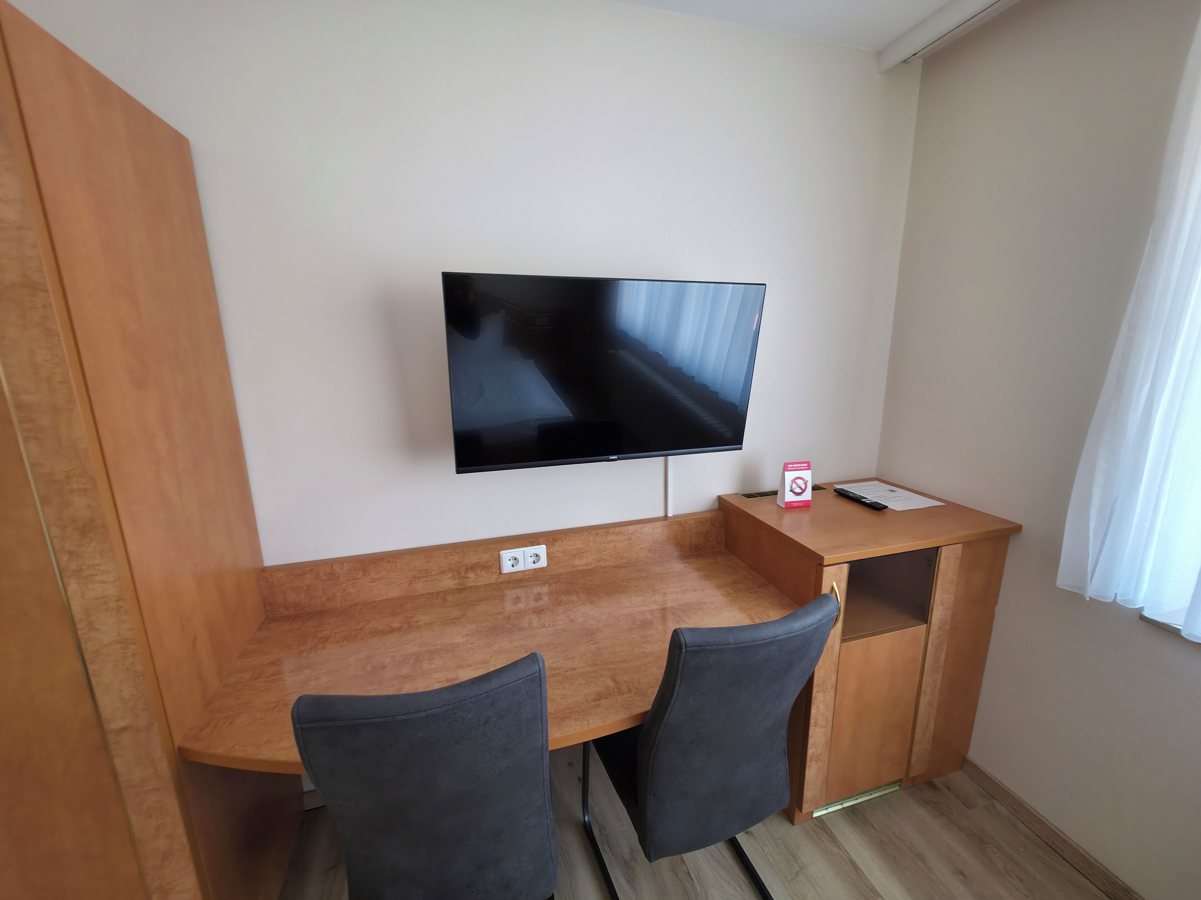 A room with a wooden desk, two chairs, a flat-screen TV mounted on the wall, a small box with a red circle and line symbol, and a window with white curtains.