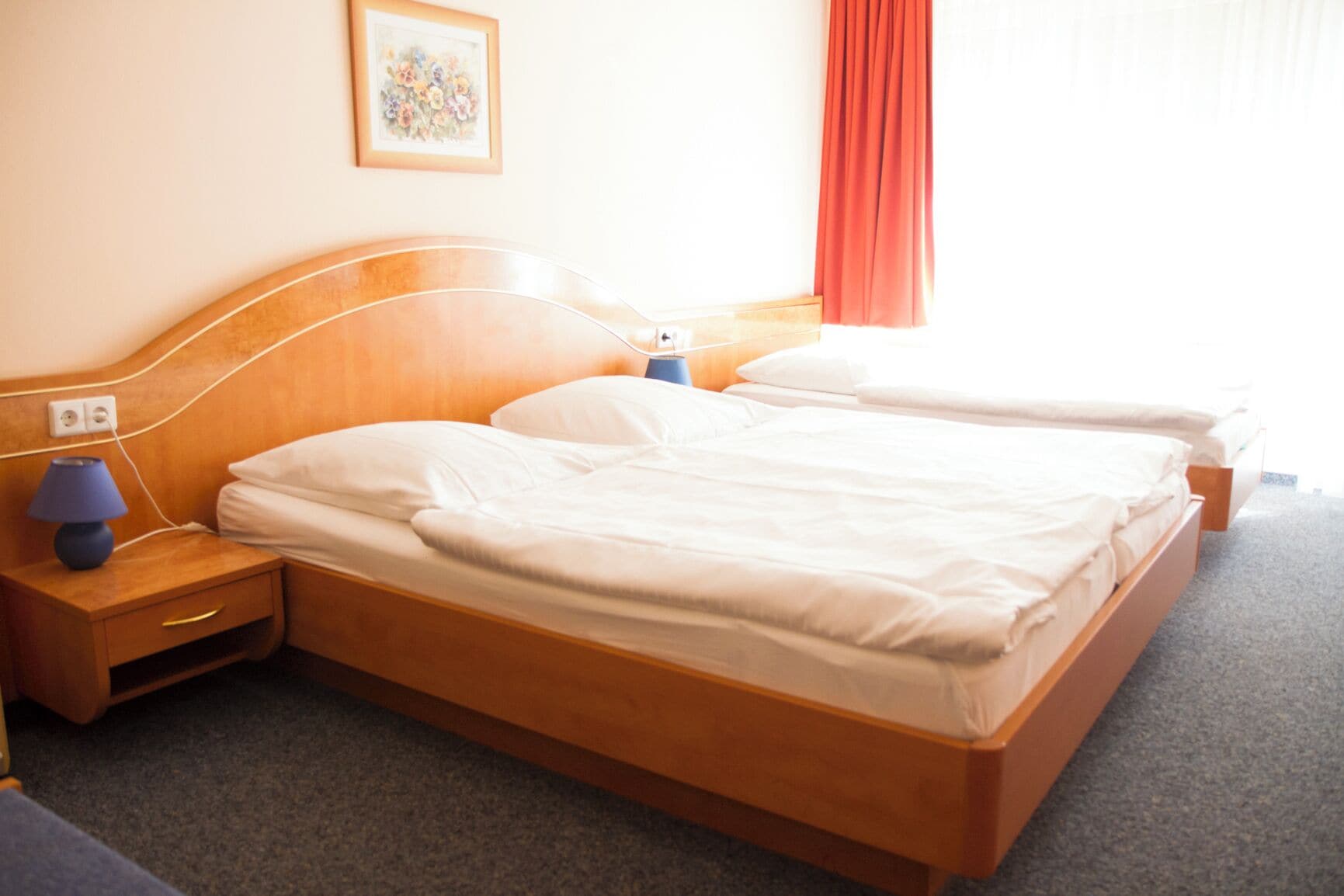 A double bed with wooden headboard, two bedside tables with lamps, a power outlet, and a framed floral artwork on the wall.