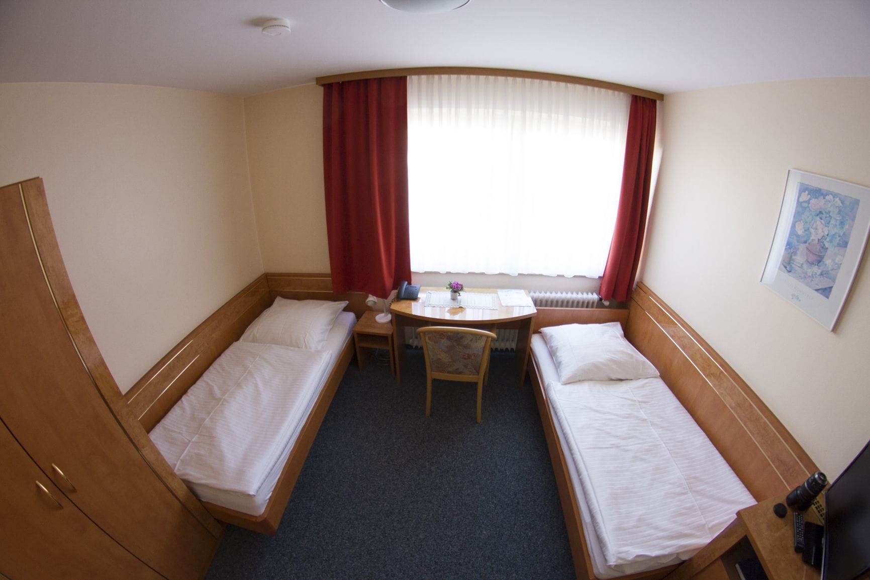 A hotel room with two single beds, a small desk with a chair, a window with red curtains, and a radiator.
