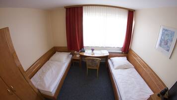 A hotel room with two single beds, a small desk with a chair, a window with red curtains, and a radiator.