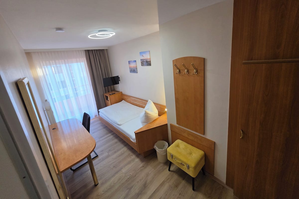 A hotel room with a bed, a wooden desk, a small yellow ottoman, a coat rack, and a trash bin.