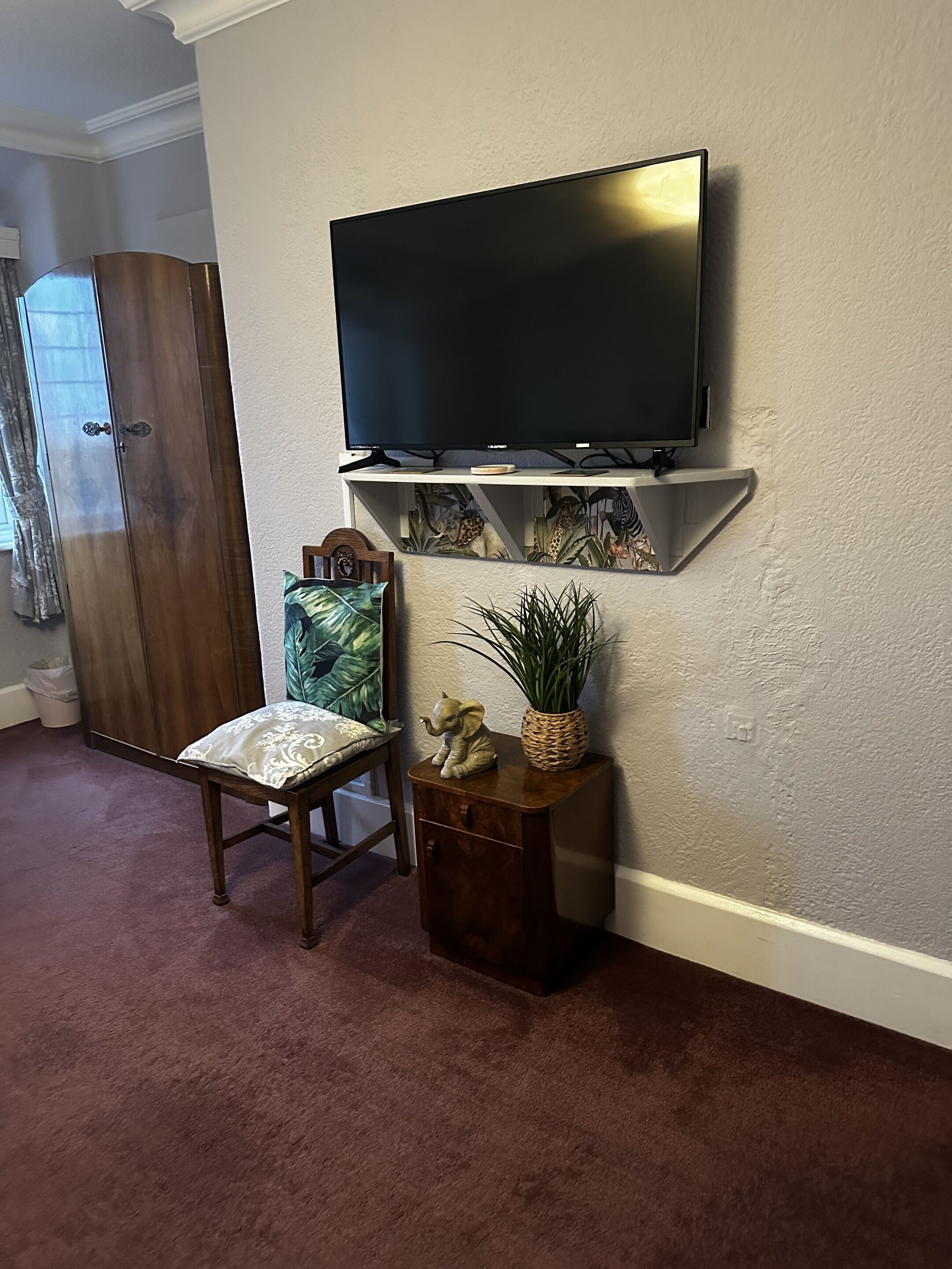 A living room with a flat-screen TV mounted on a wall, a wooden chair with a patterned cushion, a small wooden side table with a decorative elephant figurine and a potted plant, and a large wooden wardrobe.