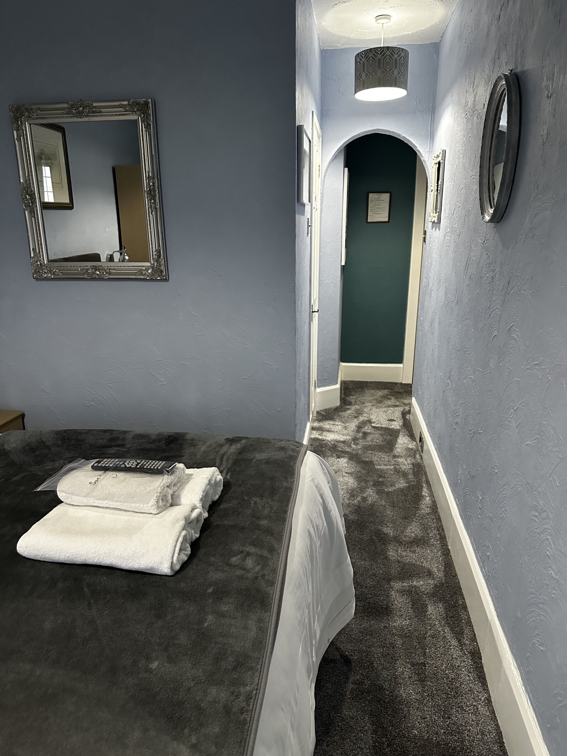 A hotel room with a bed, a large mirror, a small table with a towel and a remote control, and a hallway leading to another room.