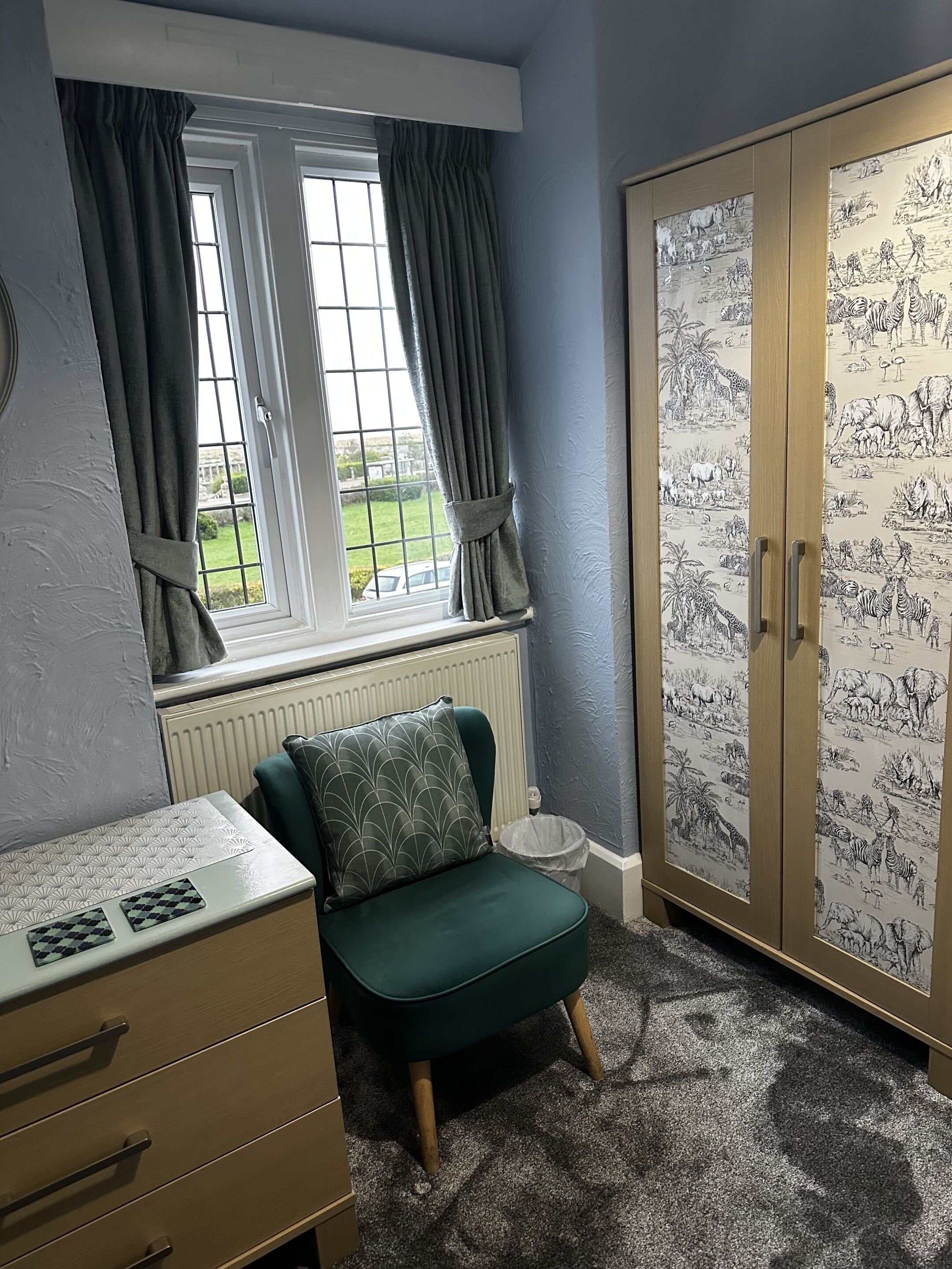 Une pièce avec une fenêtre, un radiateur, un fauteuil vert, une armoire en bois et une commode à tiroirs.