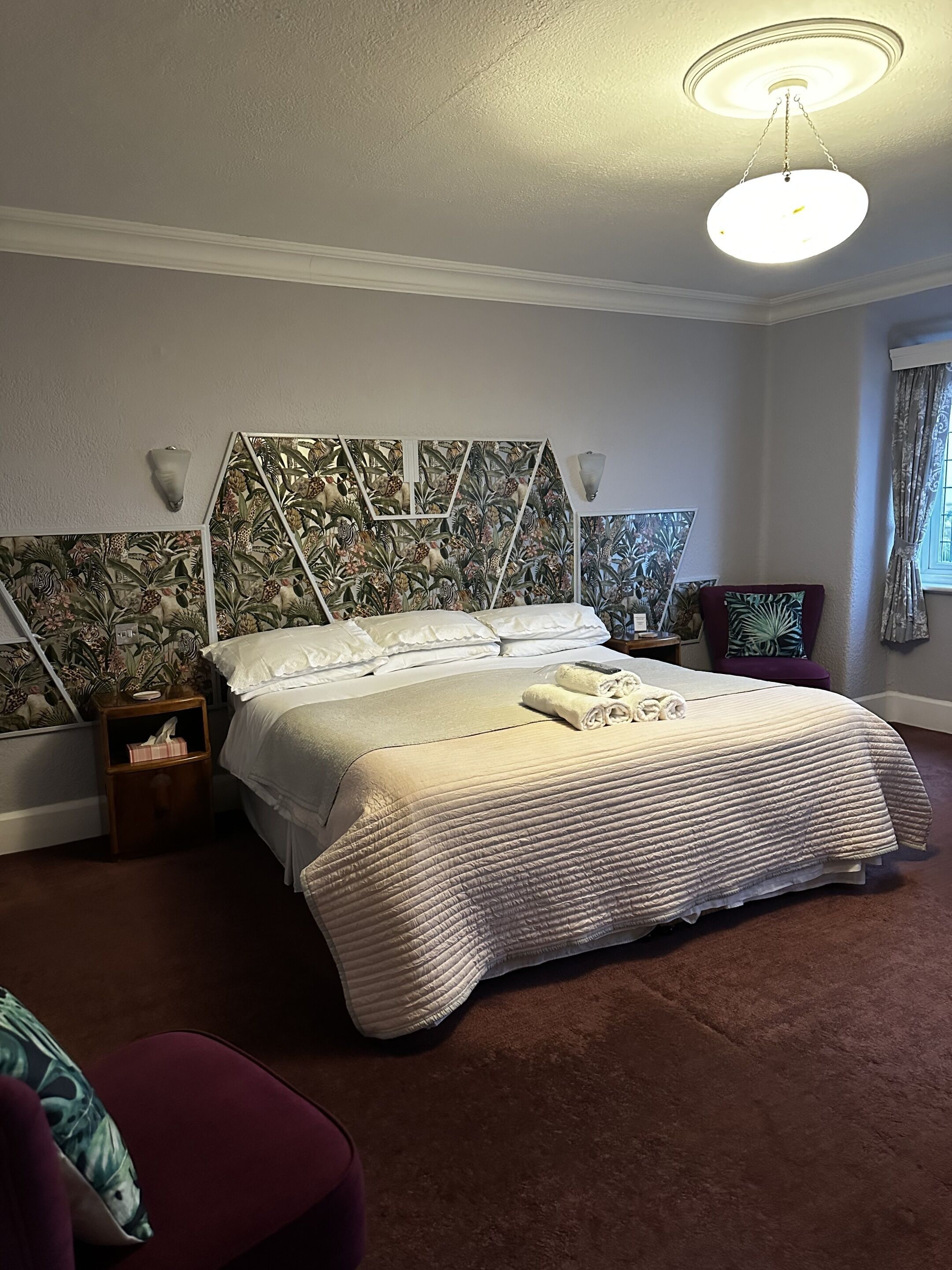 Een netjes opgemaakt bed met een wit hoofdbord, een nachtkastje met een lamp, en een muur met een tropisch behangpatroon.