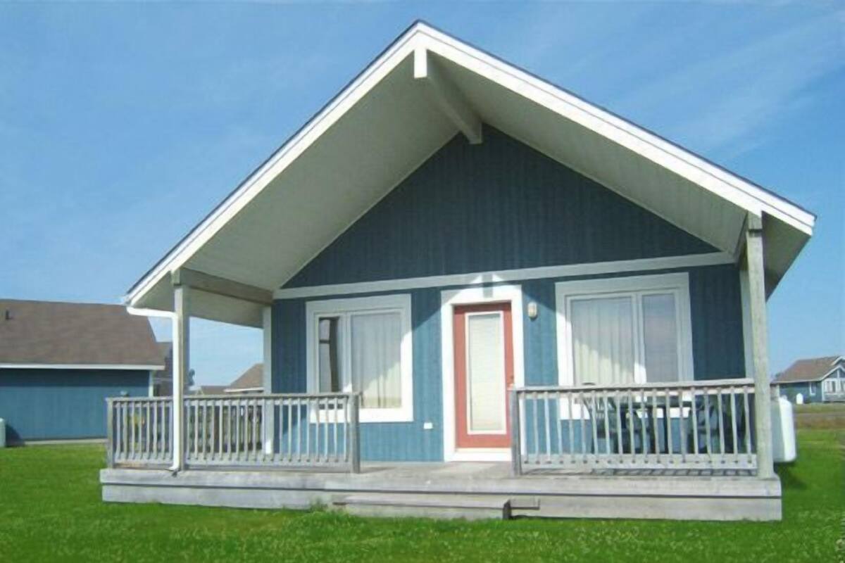 Een gelijkvloers huis met een puntige dak, een veranda met witte leuningen en een rode deur.