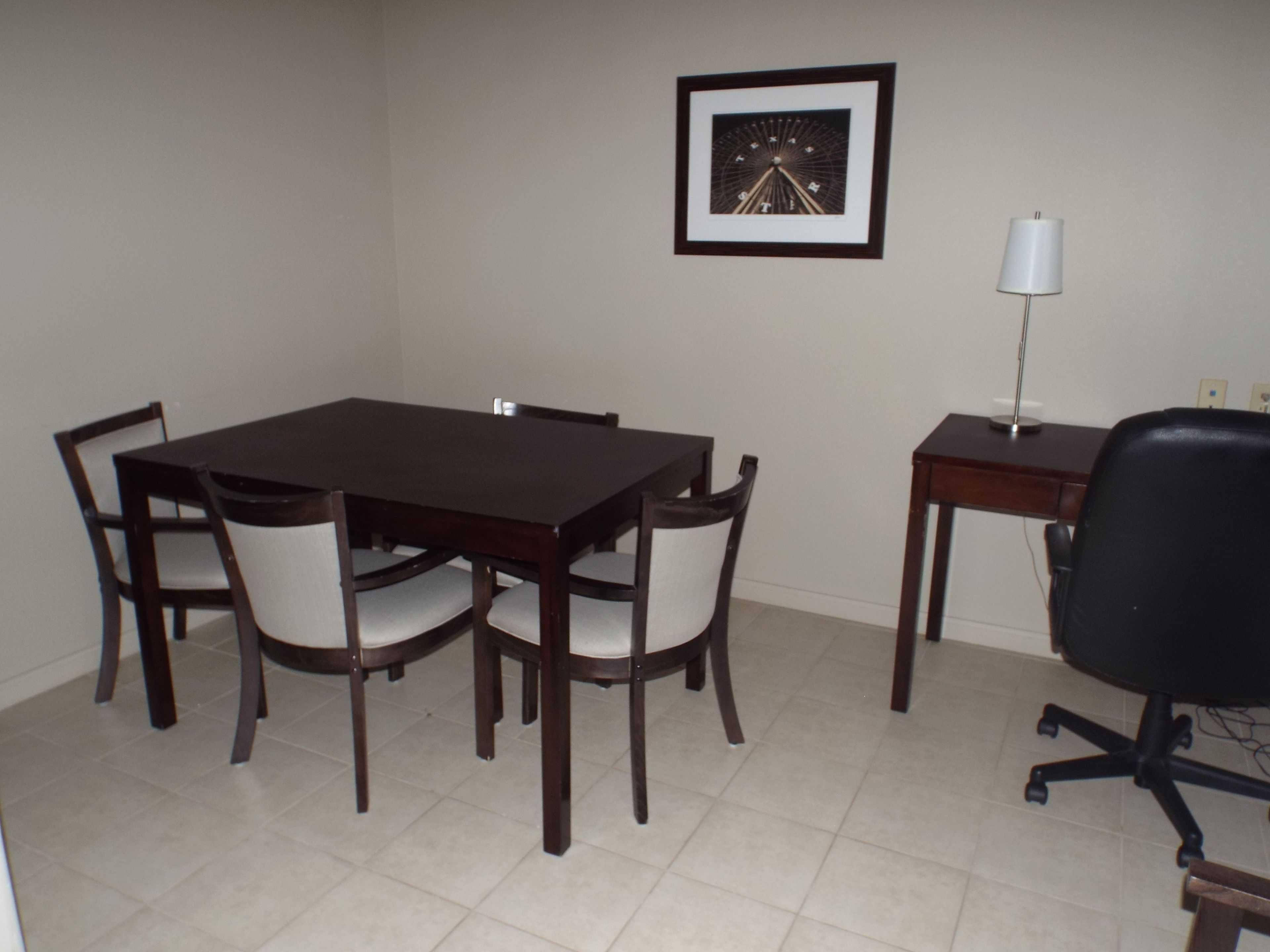 A conference room with a dark wooden table and four chairs. There is a framed picture on the wall and a desk with a lamp.