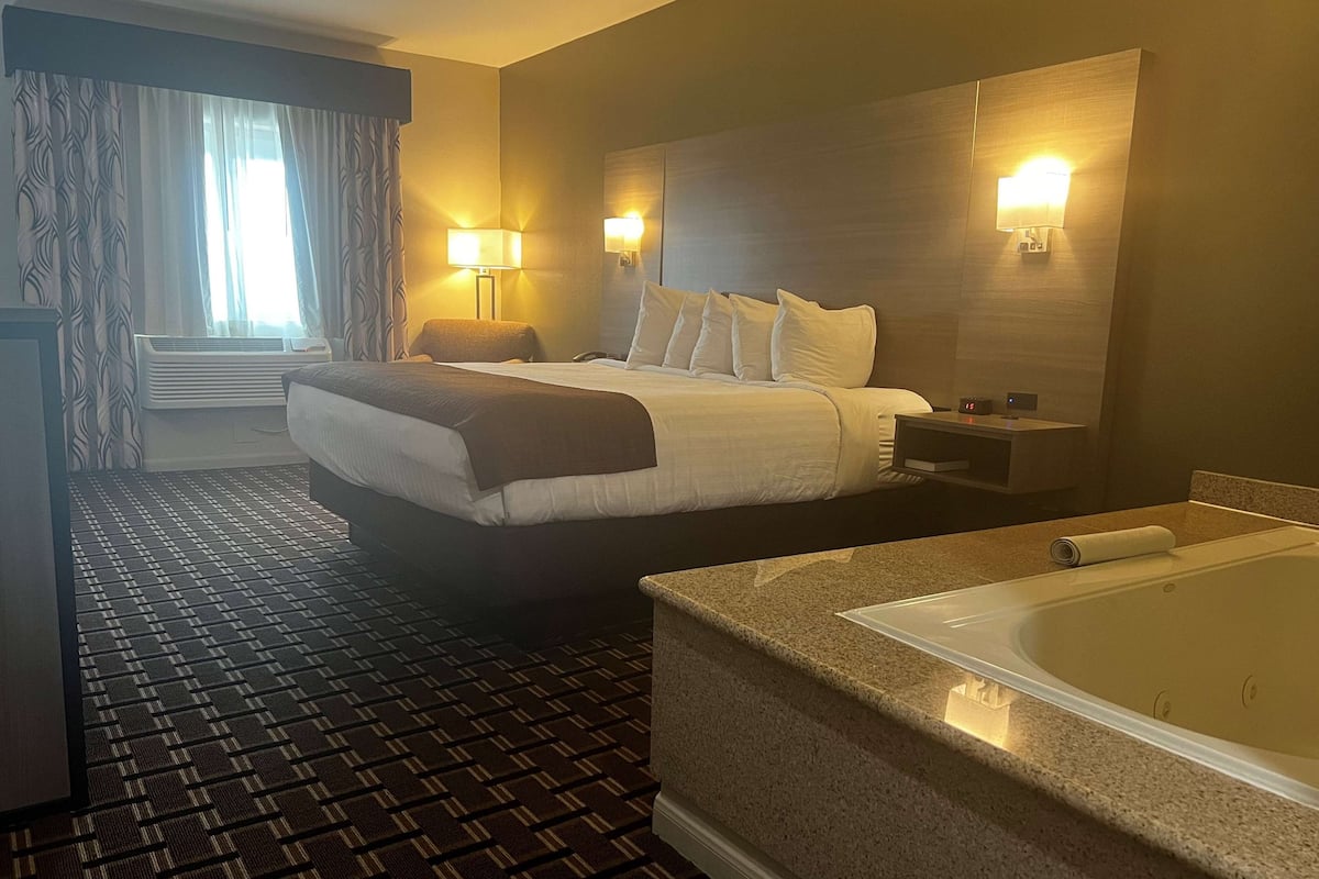 Ein Hotelzimmer mit Bett, Schreibtisch, Stuhl, Schreibtischlampe, Wandlampe, einem großen Fenster mit Vorhängen, einer Klimaanlage, einem Teppich und einer Badewanne.