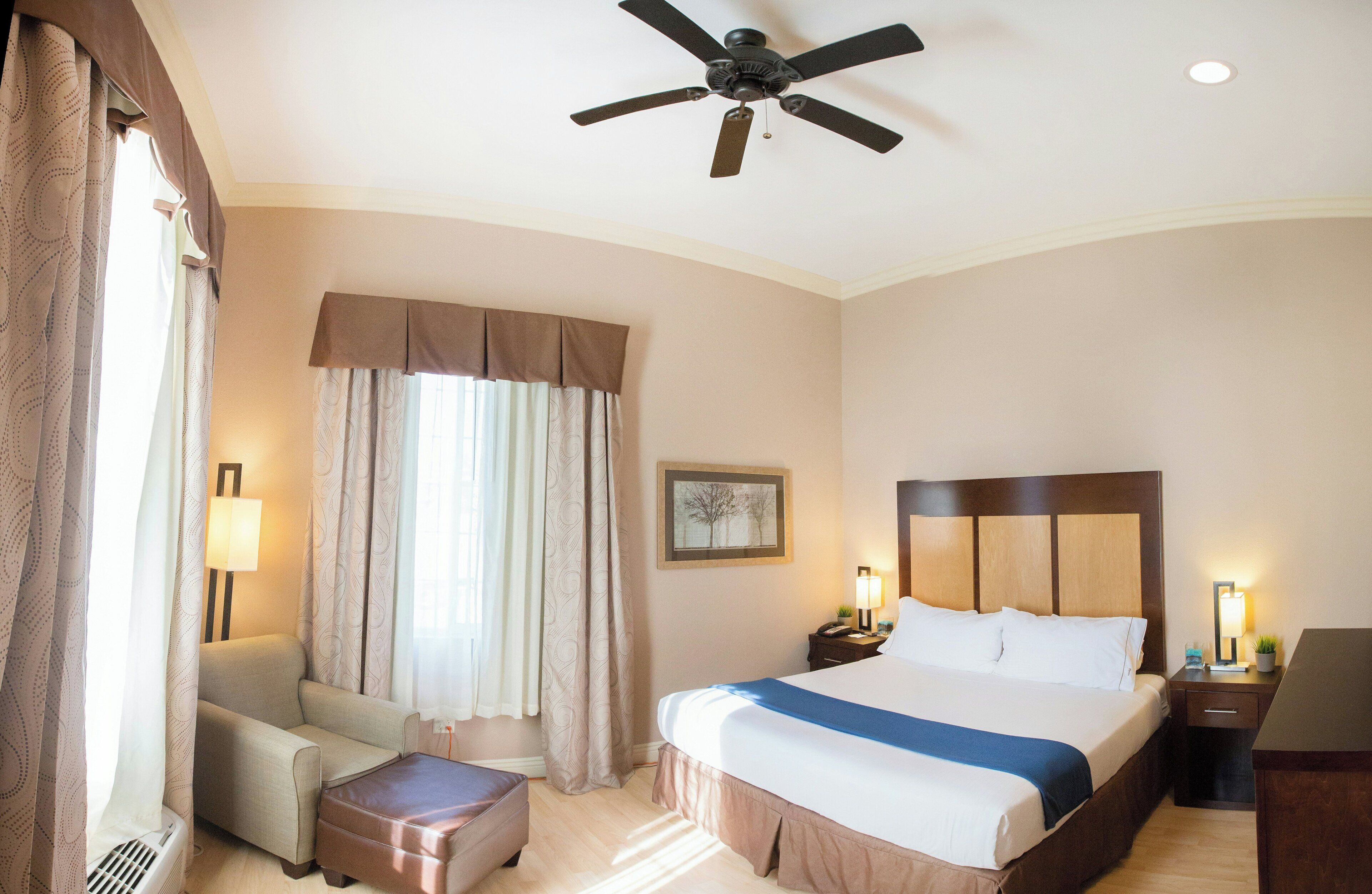 Una camera d'hotel con un letto, un comodino, una lampada, una ventola a soffitto, una finestra con tende e un quadro appeso al muro.