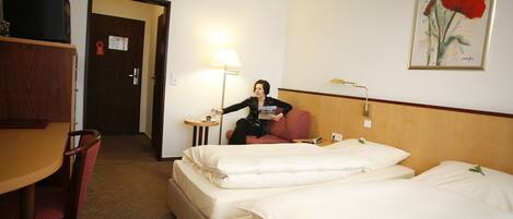 Quarto de hotel com duas camas, uma escrivaninha, uma cadeira, um abajur e uma porta.