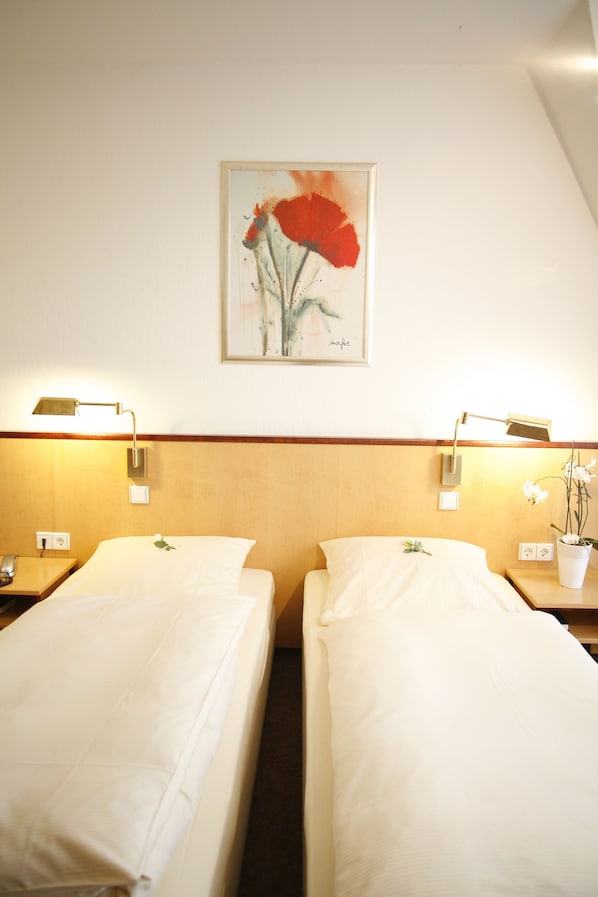 Habitación de hotel con dos camas individuales, cabecero de madera, lámparas de noche, un cuadro enmarcado de una flor y una orquídea en maceta sobre la mesita de noche.
