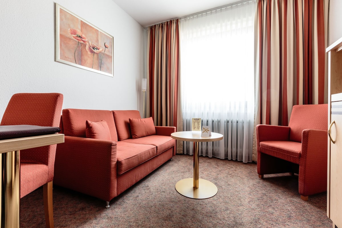 Ein Hotelzimmer mit einer roten Couch, einem kleinen runden Tisch und einem Stuhl. An der Wand hängt ein Gemälde und es gibt ein Fenster mit Vorhängen.