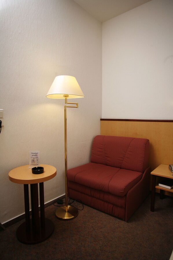 A hotel room with a red sofa, a standing floor lamp, a small round table with a lamp, and a wooden nightstand. - Hotel St. Pierre (Bad Hoenningen)