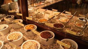 Un buffet con diversi cibi, con etichette che includono 'Herkules', 'Cornflakes', 'Choco-Crispis', 'Frittelle-Müsli', 'Toppas' e 'Krapper-Müsli'.