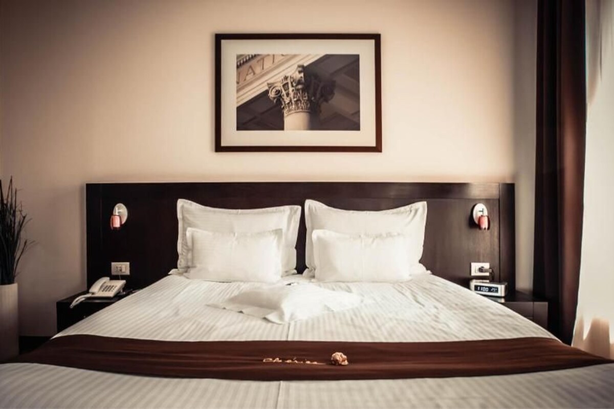 Habitación de hotel con una cama grande, dos mesitas de noche, una lámpara de noche, un cuadro enmarcado en la pared y una planta en maceta.