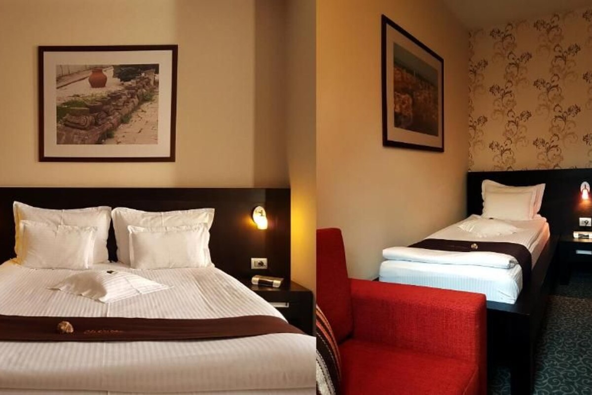Una habitación de hotel con una cama grande, una cama más pequeña, una silla roja y dos mesitas de noche con lámparas.