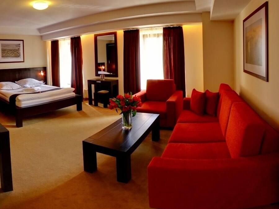 A hotel room with a bed, a red sofa, a coffee table with flowers, and a desk with a lamp.