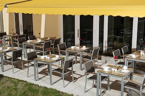 Un café al aire libre con mesas y sillas, un toldo amarillo y grandes ventanales.