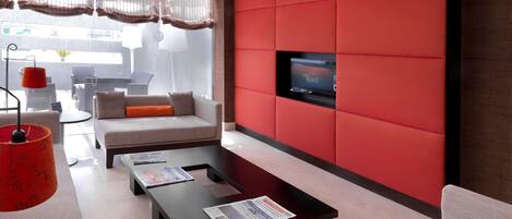 A modern living room with a red accent wall, a flat-screen TV, a sofa, a coffee table, and a window with sheer curtains.