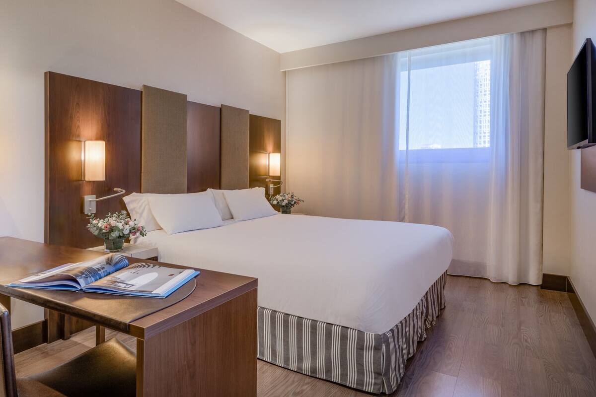 Une chambre d’hôtel avec un lit, un bureau, une lampe, une télévision et une fenêtre avec des rideaux.