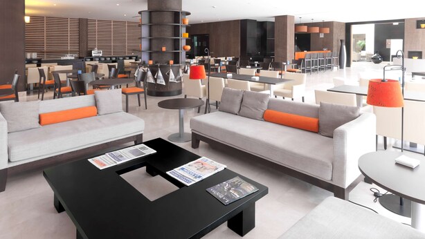 A modern hotel lobby with a seating area featuring sofas and armchairs, a coffee table with magazines, and a large cylindrical decorative piece. - NH Campo De Gibraltar (Los Barrios)