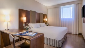 A hotel room with a large bed, a desk with a magazine, a lamp, and a window with curtains. - NH Campo De Gibraltar (Los Barrios)