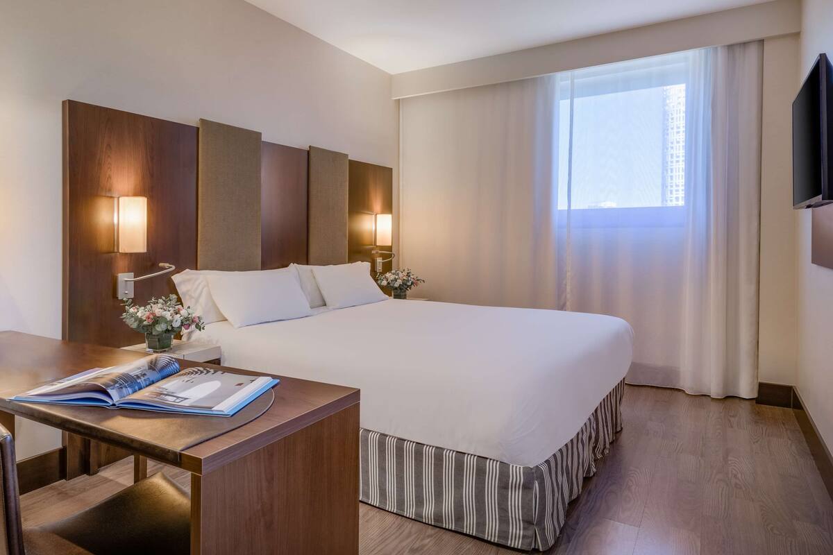 Une chambre d’hôtel avec un grand lit, un bureau avec un magazine, une lampe et une fenêtre avec des rideaux.