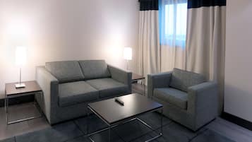 Una habitación de hotel moderna con un sofá gris, un sillón a juego, una mesa de centro de vidrio y una lámpara de pie.