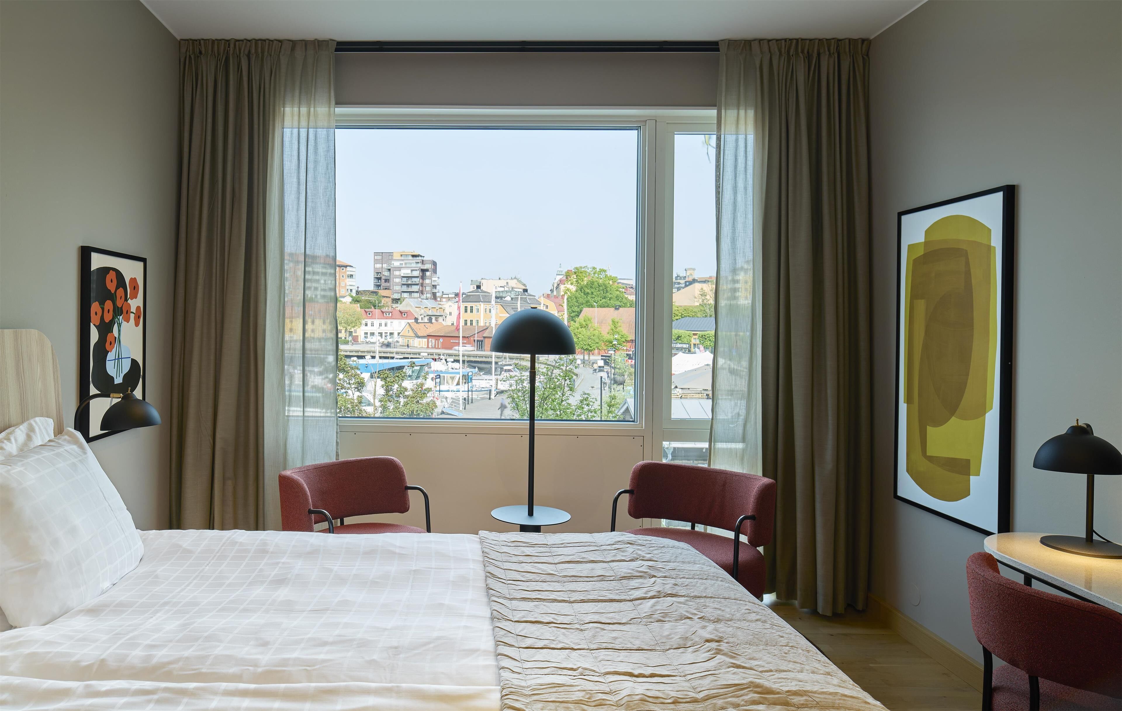 A hotel room with a large bed, two red chairs, a small table, a floor lamp, and a large window with a city view.