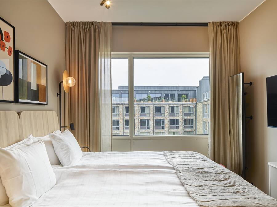 Habitación de hotel con una cama grande, un televisor de pantalla plana, un cuadro de flores y vistas a edificios a través de la ventana.
