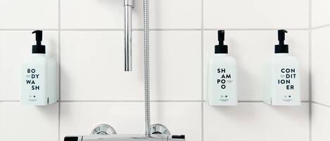 A bathroom with a chrome showerhead, a handheld showerhead, and three bottles of body wash, shampoo, and conditioner on the wall.